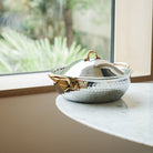 Mauviel M'ELITE B hammered 5-ply curved splayed saute pan with brass handles and lid, shown in a lifestyle kitchen setting.